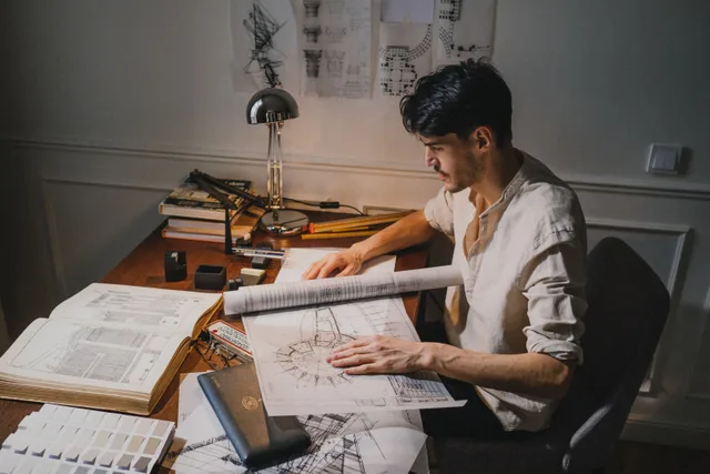 Young architect at a desk working on blueprint designs under lamp light. Recurring small home design firm problems, and the guide to fixing each