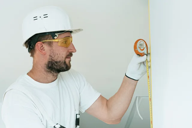 Construction worker with hard hat and goggles measuring a wall with a tape measure. Preventing small space design mistakes: tools to test and two alarms