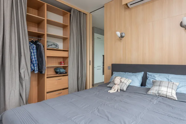Modern room interior with bed with pillows and soft rabbit toy near wooden wardrobe with curtains and shelves near clothes on hangers and entrance to kitchen in bright flat. Space saving room ideas homeowners regret, from unused fold-downs to unreachable storage