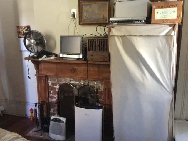 1880s House in Carrollton section of New Orleans. Bedroom wooden fireplace, on the mantle are seen an electric fan, small television, small, wooden chest. On ground in front are space heater, air filter machine. To right is a portable closet. Wildfire Smoke: Small Home Retrofit Priorities for Renters